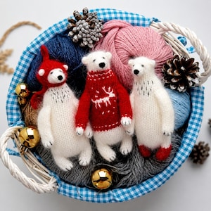 Puede incluir: Tres pequeñas figuras de osos polares tejidos en blanco, dentro de una cesta de cuadros azules y blancos con ovillos de lana, piñas y adornos dorados. Un oso lleva un gorro rojo, otro un suéter rojo con un ciervo blanco y otro pies rojos.