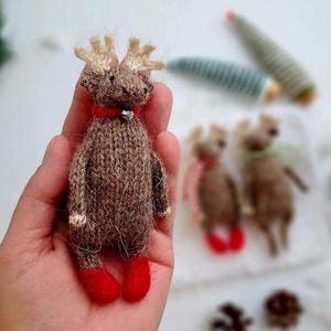 May include: A handmade brown knitted reindeer doll with red felt feet, a red collar, and small antlers. The reindeer is held in a hand, with other similar dolls in the background. The doll has a small silver bell on its collar.