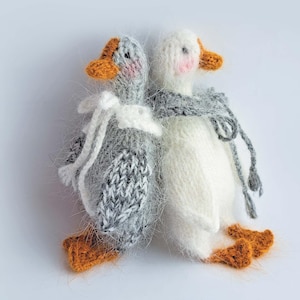 Puede incluir: Dos muñecos de ganso tejidos. Uno es gris con un lazo blanco, el otro es blanco con una bufanda gris. Ambos tienen picos y patas naranjas, y mejillas rosadas. Los muñecos están hechos a mano y tienen una textura suave y esponjosa.