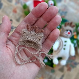 May include: A miniature, hand-knitted hat in shades of tan and brown, featuring small antlers. The hat has delicate ties and is held in a person's hand, with a blurred background showing a small snowman figure.