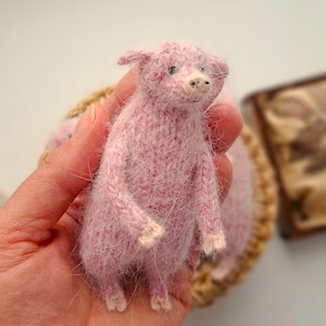 Puede incluir: Una figura de cerdo rosa en miniatura hecha a mano. El cerdo está hecho de un material suave y texturizado, con costuras y detalles visibles. Tiene pequeñas pezuñas blancas y una cara detallada. El cerdo se sostiene en una mano, con un fondo borroso.