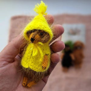 Puede incluir: Un osito de peluche en miniatura hecho a mano que lleva un impermeable con capucha amarillo brillante. El oso es marrón con patas naranjas y una nariz negra. El impermeable tiene una capucha a juego con una borla y un cierre de lazo. Otros dos osos en miniatura están borrosos en el fondo.