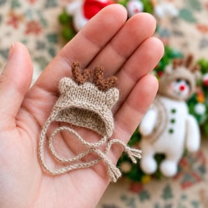 Puede incluir: Un gorro de punto en miniatura de color beige con pequeñas astas marrones y orejas redondeadas. El gorro incluye dos finas cintas trenzadas que se extienden desde los lados, diseñadas para sujetar el accesorio. La textura es suave.