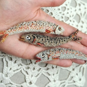 May include: Three miniature crocheted fish in shades of grey, brown, and orange are displayed. The fish have detailed features, including fins and eyes. The fish are held in a hand, showcasing the intricate craftsmanship of the handmade items.