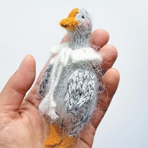 May include: A handmade miniature goose doll with a gray body, white and black patterned wings, and an orange beak and feet. The doll has a white collar and is held in a hand.