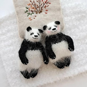 May include: Two handmade panda bear stuffed animals with black and white fur. The pandas have small black eyes and pink cheeks. They are positioned in front of a beige embroidered fabric with a floral design.