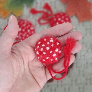 Puede incluir: Primer plano de un adorno en forma de hongo rojo con puntos blancos, sostenido en una mano. El adorno tiene una textura difusa y un hilo rojo adjunto. Otros adornos similares son visibles en el fondo.