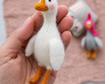 May include: A small, hand-knitted white goose toy with a fuzzy texture, featuring a bright orange beak, orange feet, and small pink circular patches on its cheeks. The goose has a single black bead eye visible on the side of its head.