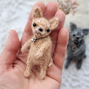 May include: A small, tan-colored knitted Chihuahua dog with black eyes and a silver bell collar. The handmade dog has a detailed texture and is held in a person's hand. A second, gray knitted dog is in the background.