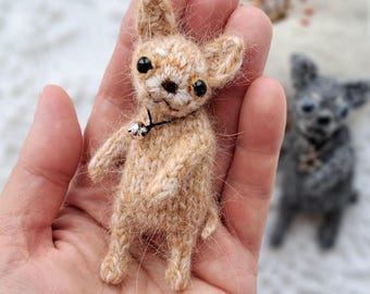 May include: A miniature, hand-knitted Chihuahua dog in tan with black eyes and a silver bell collar. The dog is held in a hand, showcasing its detailed texture. A second, grey knitted dog is visible in the background.