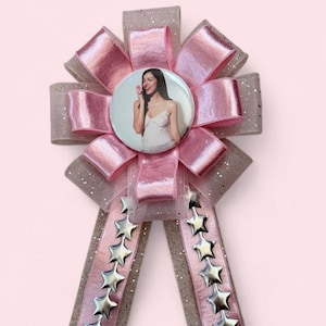 Op de afbeelding: Een decoratieve rozet met een ronde speld waarop een foto van een persoon met een bloem staat. De rozet is gemaakt van gelaagd roze satijn en beige glitterlint, met twee lange uiteinden die zijn versierd met een verticale rij zilveren sterren.