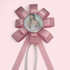 May include: A pink rosette badge featuring a circular button with a photograph of a person in a white top. The rosette is constructed from layered glittery and satin pink ribbons, with two long, light-coloured ribbons hanging down, each printed with a vertical row of small pink hearts.