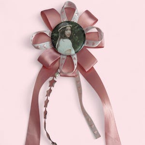 May include: A decorative rosette badge featuring a circular pin with a photographic portrait of a person wearing a floral crown. The badge is surrounded by layered dusty rose satin ribbons and a thinner ribbon with small heart patterns.
