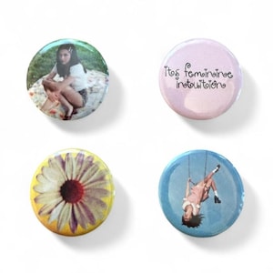 May include: Four circular pin-back badges. One features a person sitting on a patterned blanket, one is light purple with the text 'it's feminine intuition', one shows a yellow and purple daisy, and one depicts a person on a swing against a blue sky.