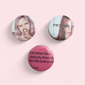 May include: Three circular pin-back badges. The top left badge features a person with pink hair biting a finger. The top right badge shows a person with red hair and the text PRIMA. The bottom badge is pink with the text I'd rather be a nobody than to be like everyone and the Spotify logo.