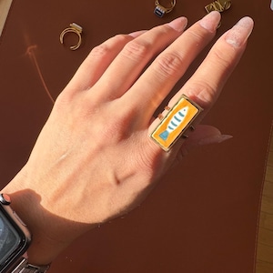 May include: Gold-coloured rectangular ring with a hand-painted fish design. The fish is blue and white on an orange background. The ring is worn on a finger, with other rings visible in the background.
