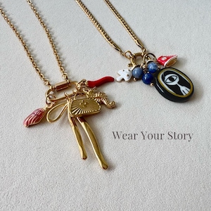 May include: Two gold-toned chain necklaces featuring assorted charms. One necklace has a red chili, a shell, and a gold fan. The other has a white mushroom, blue beads, a red shoe, and a black oval pendant with an eye graphic. Text reads Wear Your Story.