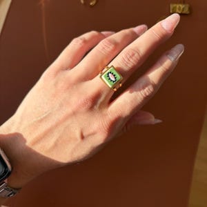 May include: Gold ring with a square, light green enamel face, featuring a white oval and pink and black details. The ring is displayed on a finger, highlighting its design and colour contrast.