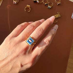 May include: A gold ring with a square, pale blue face featuring an eye design with pink and white details. Other rings with various designs are visible in the background.
