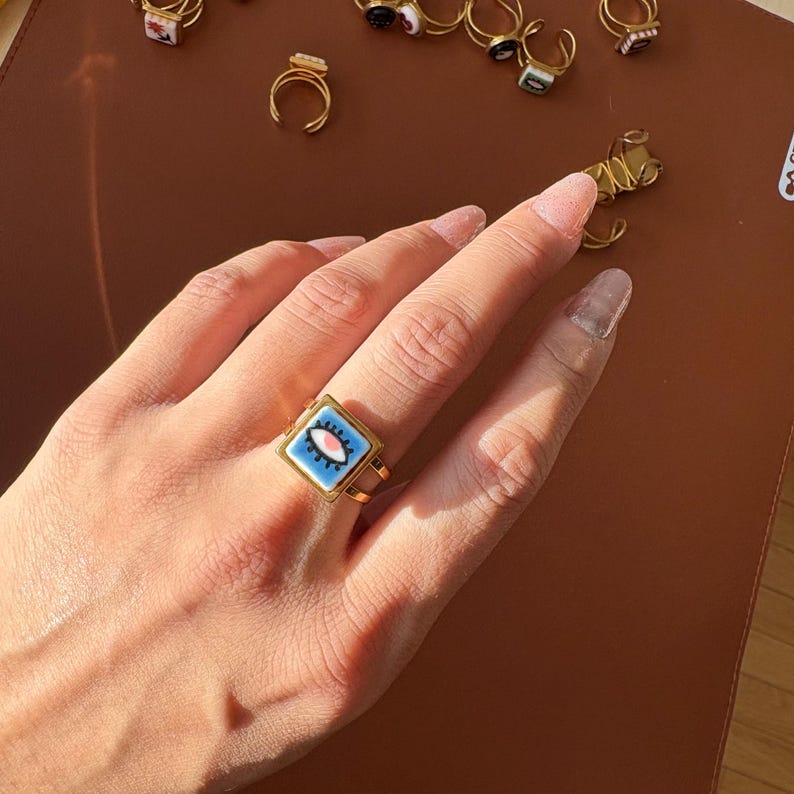 May include: A gold ring with a square, pale blue face featuring an eye design with pink and white details. Other rings with various designs are visible in the background.