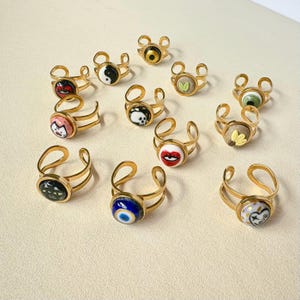 May include: A selection of gold-toned adjustable rings, each with a unique design. The rings have decorative, circular tops featuring images such as a skull, lips, and an eye. The rings are presented on a light-coloured surface.