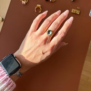 May include: A gold-coloured ring with a black and white yin-yang symbol. The ring has a circular design with a raised bezel. Other rings are visible in the background. The ring is on a brown surface.