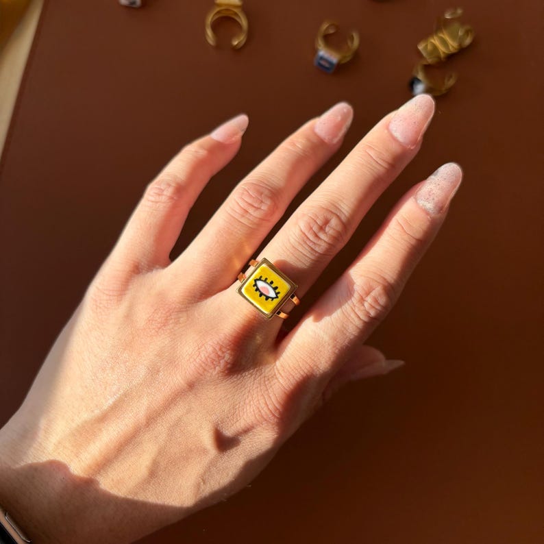 May include: A gold ring with a square, yellow face featuring a white and pink eye design. The ring is on a hand with long, manicured nails. Other gold rings are visible in the background.