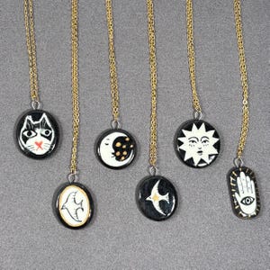 May include: A collection of gold chain necklaces with black and white pendant charms. The charms feature various designs, including a cat face, a crescent moon, a sun with a face, a bird, a flying bird, and a hand with an eye.