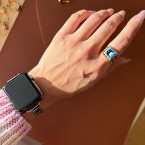 May include: A gold ring with a square, blue and white dolphin design. A silver smartwatch with a black screen is on a wrist. The background is a brown surface. The ring is on a hand.