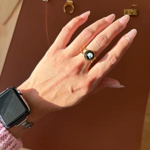 May include: A gold ring featuring a black and white skull motif. The ring, worn on a finger, has a circular black background with a white skull at its centre. The band of the ring is thick and gold.