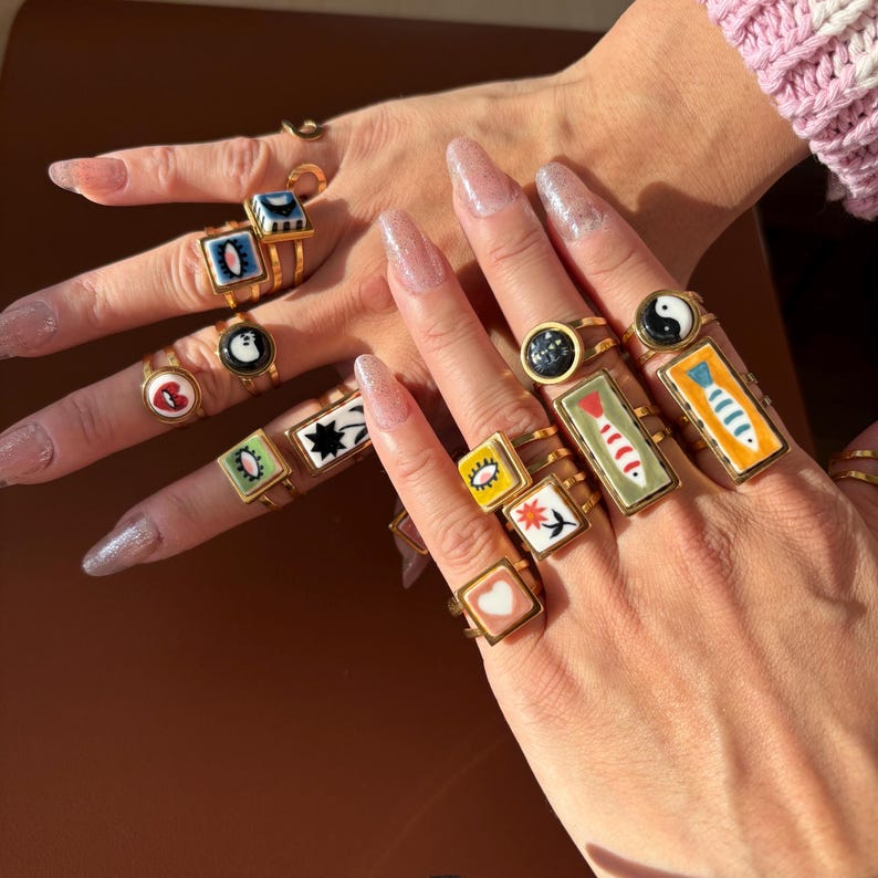 May include: A selection of gold rings, each featuring a unique, colourful design. The rings showcase motifs such as a heart, fish, and yin-yang symbol. The rings are displayed on two hands with long, manicured nails.