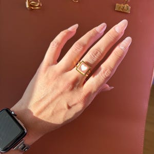 May include: A gold ring with a square face and a white heart design. The ring is worn on a hand with long, manicured nails. Other gold accessories are visible in the background.