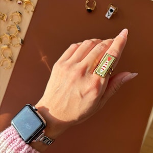 May include: A gold ring with a rectangular, pale green face featuring a red triangle and white curved lines. The ring is on a hand with a smartwatch. Other gold rings are visible in the background.
