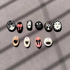 May include: A selection of ten oval and circular pendants with diverse designs. The pendants showcase a black cat, a pink bow, a hand, an eye, a sun, and a heart. The pendants are in black, pink, and cream colours.