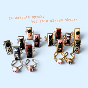 May include: A collection of cat-themed rings with rectangular and round faces. The rings feature various cat designs, including black cats, cats with coffee cups, and a cat skull. The text "It doesn't speak, but it's always there" is visible.