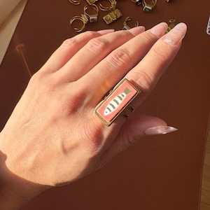 May include: A gold-coloured ring with a rectangular, coral-coloured face. The face features a white and black fish design. The ring is worn on a hand with long, manicured nails. Other rings are visible in the background.