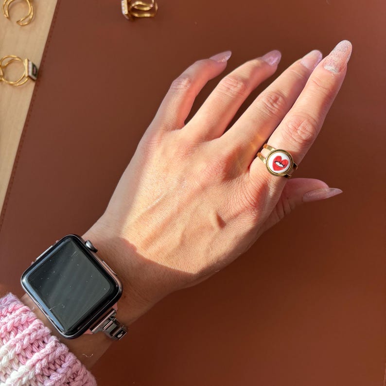 May include: A gold ring featuring a red heart and lips design on a white backdrop. The ring is worn on a hand, with a silver smartwatch on the wrist. Other gold rings are visible in the background.