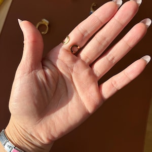May include: A hand showing several gold rings, including a small, open circle ring and a ring with a white stone. The rings are set against a brown background, with the hand's skin tone visible.
