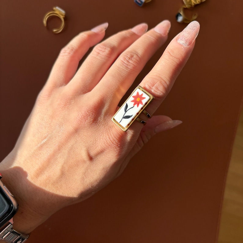 May include: A rectangular gold ring with a white enamel face featuring a red flower with black stem and leaves. The ring has a double band design and is worn on a finger.