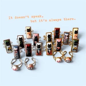 Könnte beinhalten: Eine Sammlung verstellbarer Metallringe mit rechteckigen und runden Cabochons, die verschiedene Katzenillustrationen zeigen. Der Text "It doesn't speak, but it's always there." steht über den Ringen, die Katzen in verschiedenen Outfits darstellen.