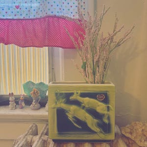 May include: A square, light green ceramic vase with a black background and yellow deer design. The vase holds pink and green dried flowers. The vase is on a wooden surface near a window with a pink and white polka dot curtain.