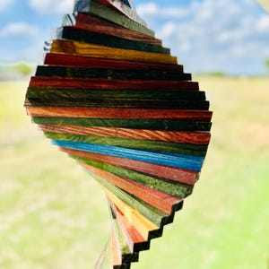 May include: A colorful wooden wind spinner with a spiral design. The wind spinner is made of various wood pieces in shades of red, green, blue, yellow, and brown. It hangs from a thin wire against a blurred background of blue sky and green grass.