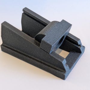 May include: A black, 3D-printed object with a rectangular base and a central, elevated component. The object has a textured surface and appears to be a mechanical part or a tool component. The design includes slots and a central opening.