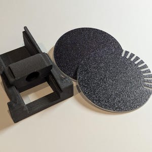 May include: A black 3D printed bracket with a central opening and two circular black textured discs, one of which features a series of small radial notches along its outer edge, designed for mechanical assembly or mounting purposes.