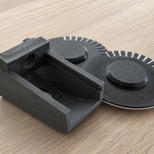 May include: A black 3D printed plastic component featuring a rectangular bracket with a hollow centre and a circular opening, attached to two adjacent circular discs with white radial markings along their outer edges and a textured surface finish.