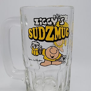 May include: A clear glass beer mug with a yellow handle. The mug has a cartoon image of a character named "Ziggy" holding a beer and a dog. The text "Ziggy's SudzMug" is printed on the mug.