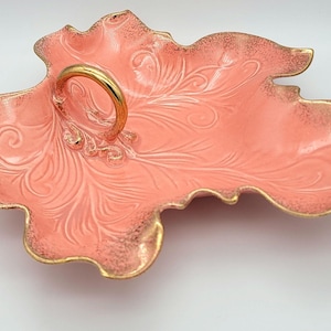 May include: A pink ceramic serving dish with a gold rim and a gold handle. The dish has a leaf-like shape and a decorative pattern.