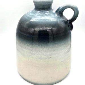 May include: A ceramic jug with a handle, featuring a gradient of colors. The top is a light blue, transitioning to dark blue and then to a creamy white at the base. The jug has a rounded body and a small opening.