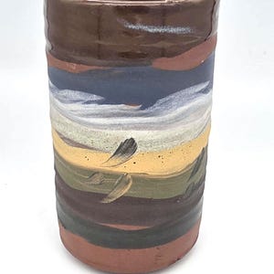 May include: A cylindrical ceramic mug with a hand-painted design. The mug features horizontal bands of color, including brown, blue, yellow, and green. The design includes brushstrokes. The mug is suitable for hot or cold beverages.