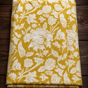 Op de afbeelding: Een gevouwen stuk stof met een mosterdgele achtergrond en een herhalende witte bloemen- en plantenprint. Het patroon bestaat uit diverse gestileerde bloemen, bladeren en stengels die in een dicht, doorlopend ontwerp over het oppervlak zijn verdeeld.